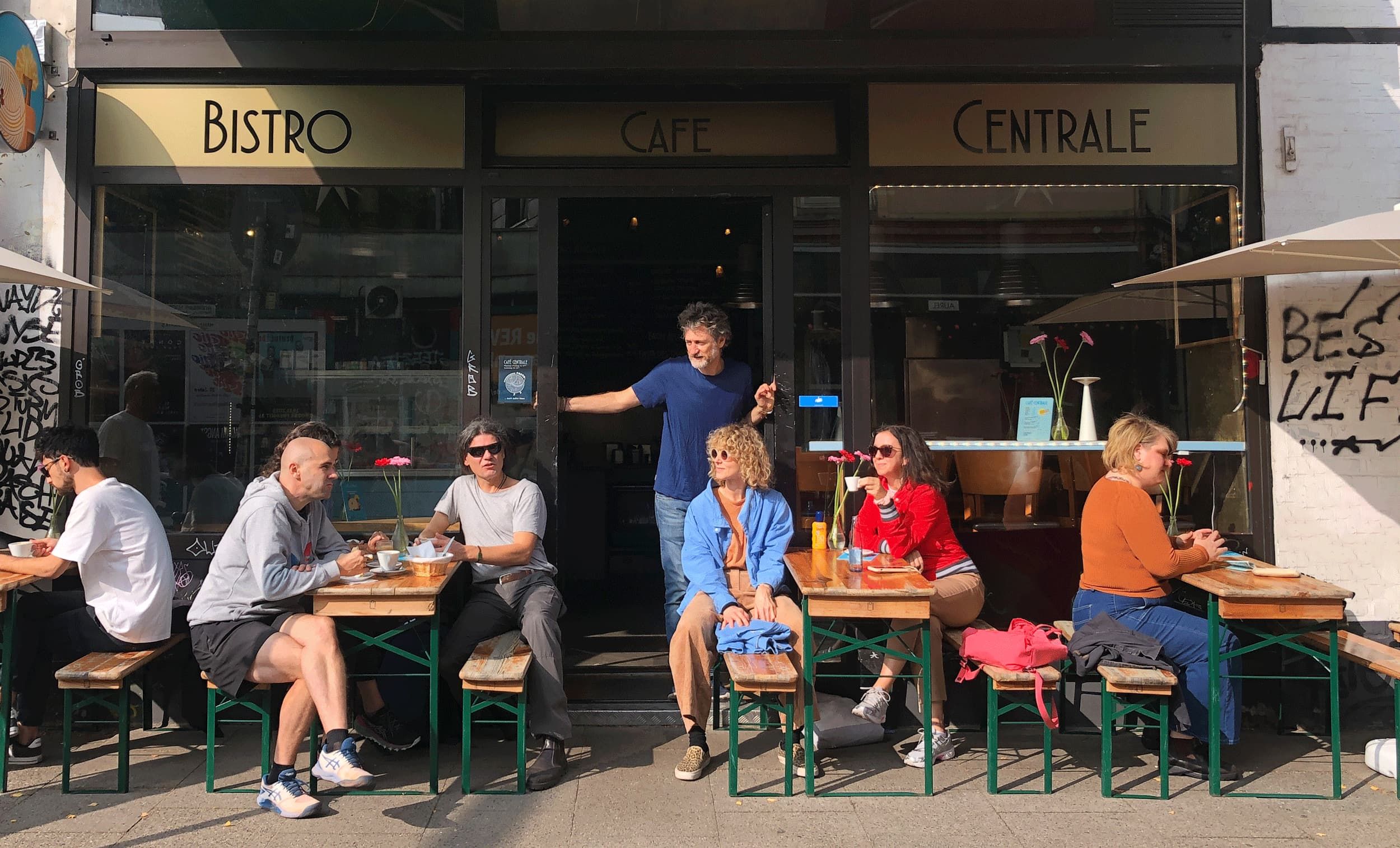 Ein Bild von Gästen, die vor dem Café sitzen, und Romeo, der im Eingang steht und das Treiben beobachtet.